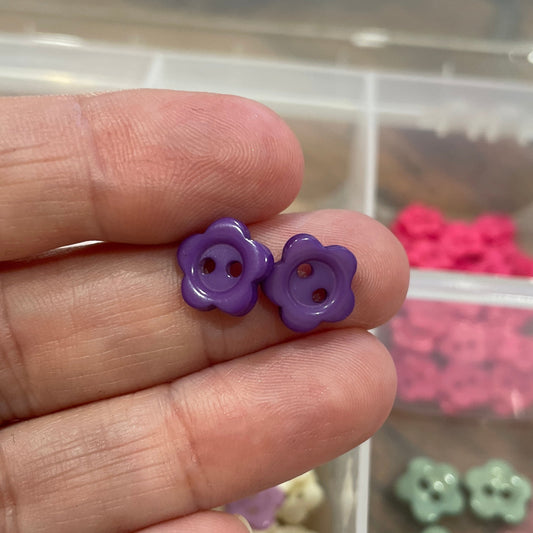 Purple flower-shaped button held between fingers with a container of assorted buttons in the background.