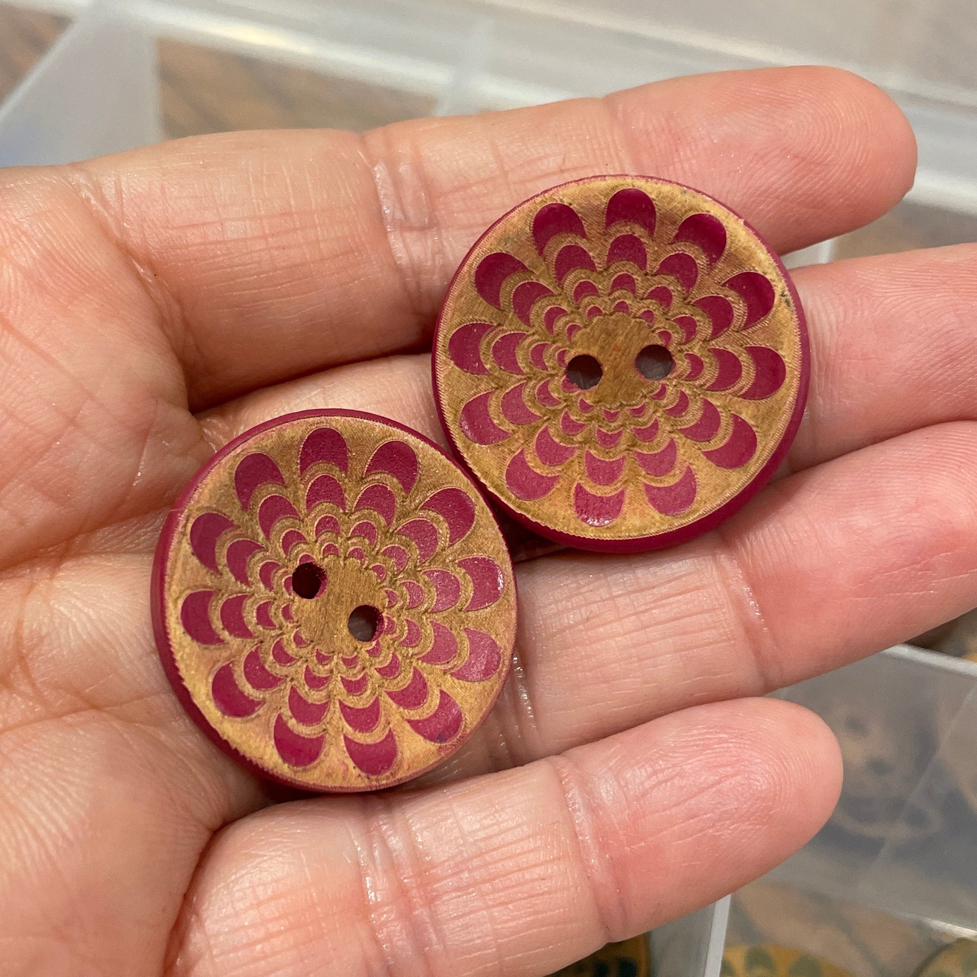 Hand holding two wooden buttons with intricate designs, with a container of similar buttons in the background.
