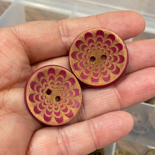 Hand holding two wooden buttons with intricate designs, with a container of similar buttons in the background.