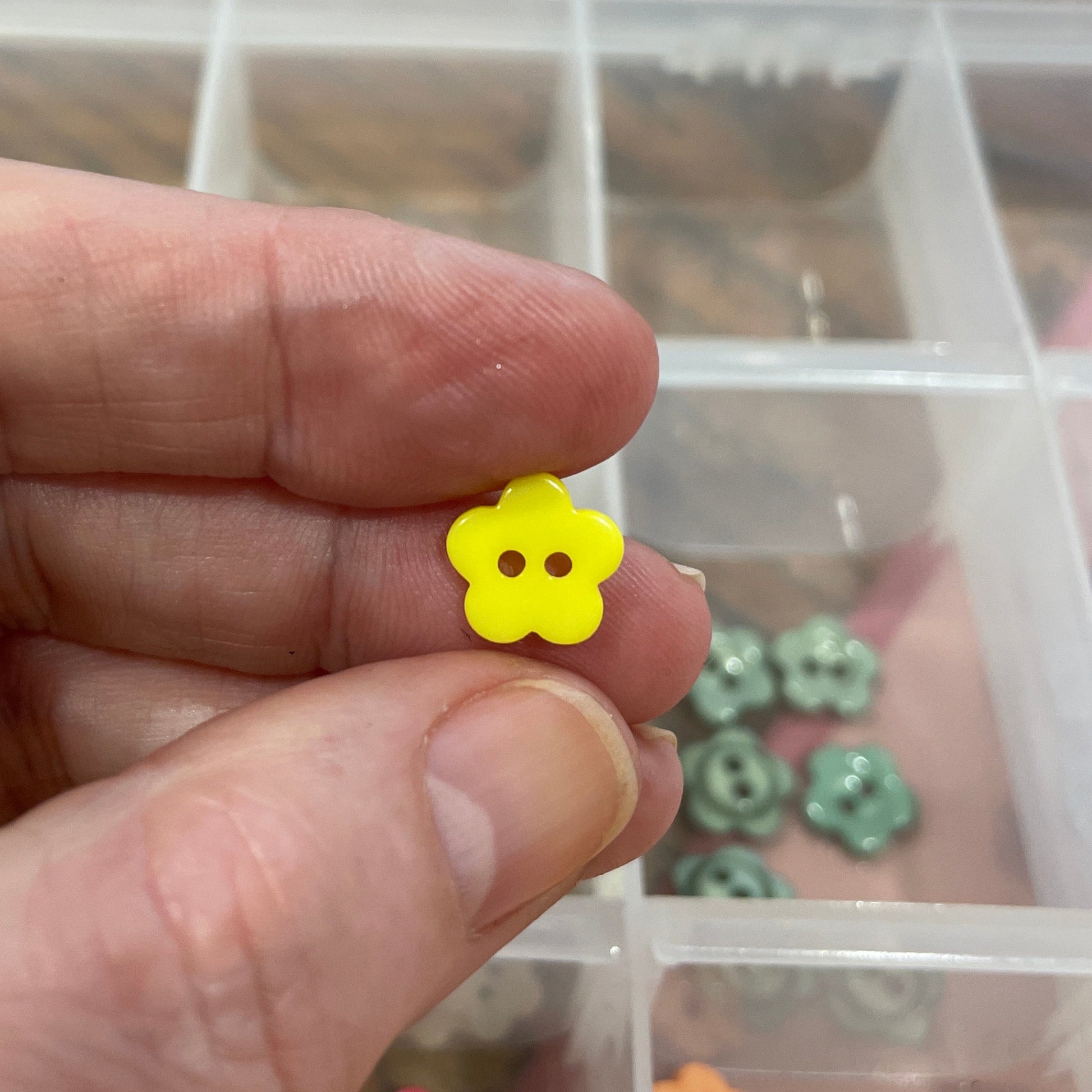 Hand holding a small yellow flower-shaped object with a clear container of colorful items in the background.