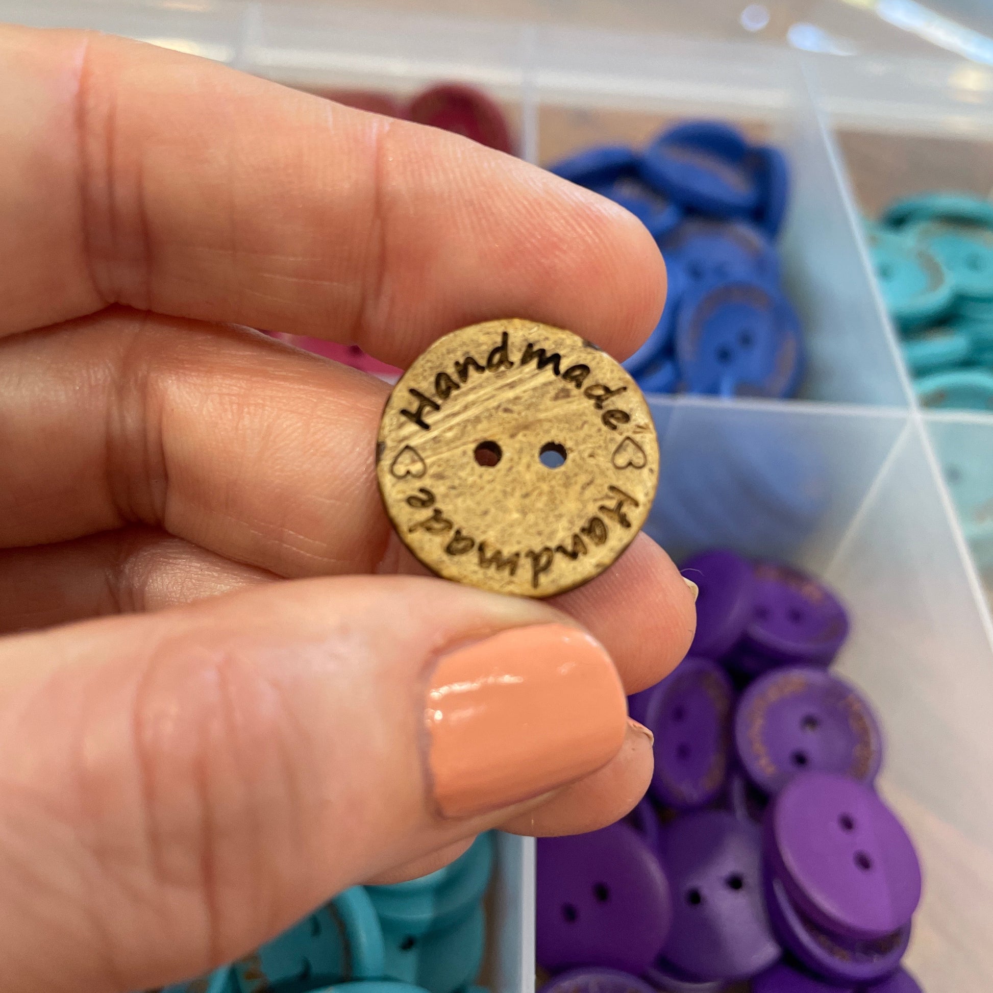Button with 'Handmade' engraving held by a hand, with button assortment in the background.