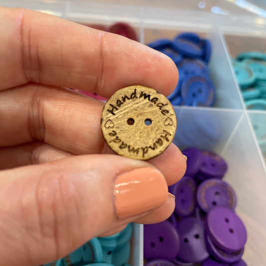 Button with 'Handmade' engraving held by a hand, with button assortment in the background.