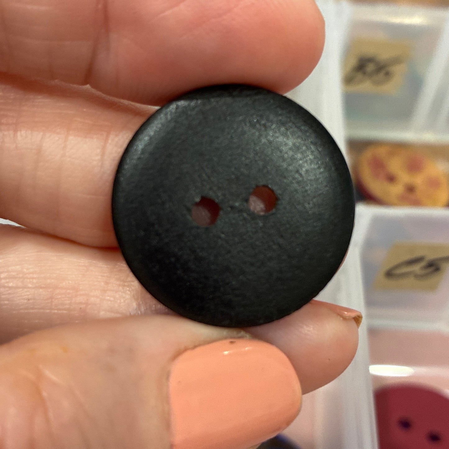 Black button held between two fingers with a background of colorful buttons in a case.