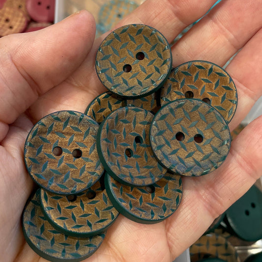 Hand holding green woven patterned buttons with a container of buttons in the background.
