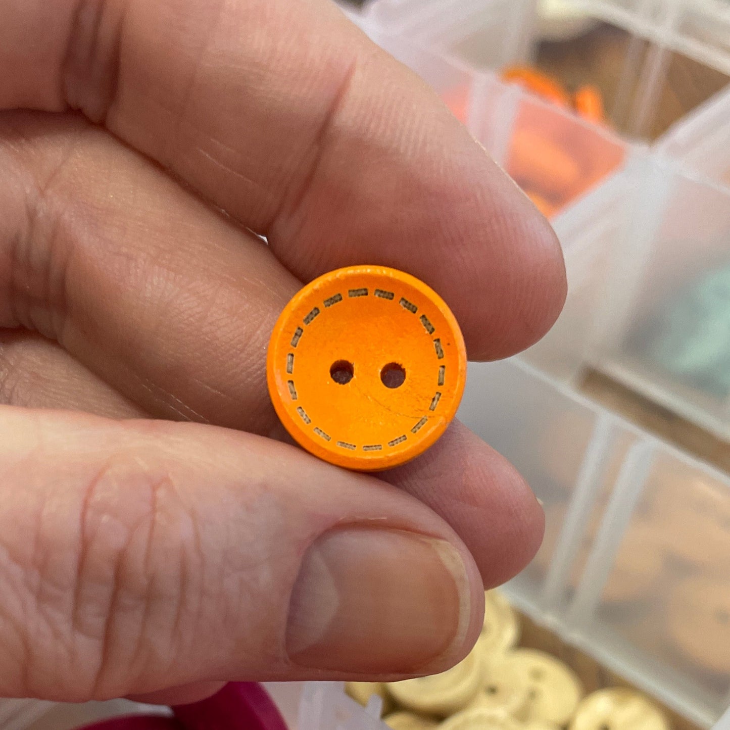 Orange button held by a hand with a container of colorful buttons in the background.