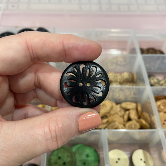 Hand holding a black decorative button with a container of assorted buttons in the background.