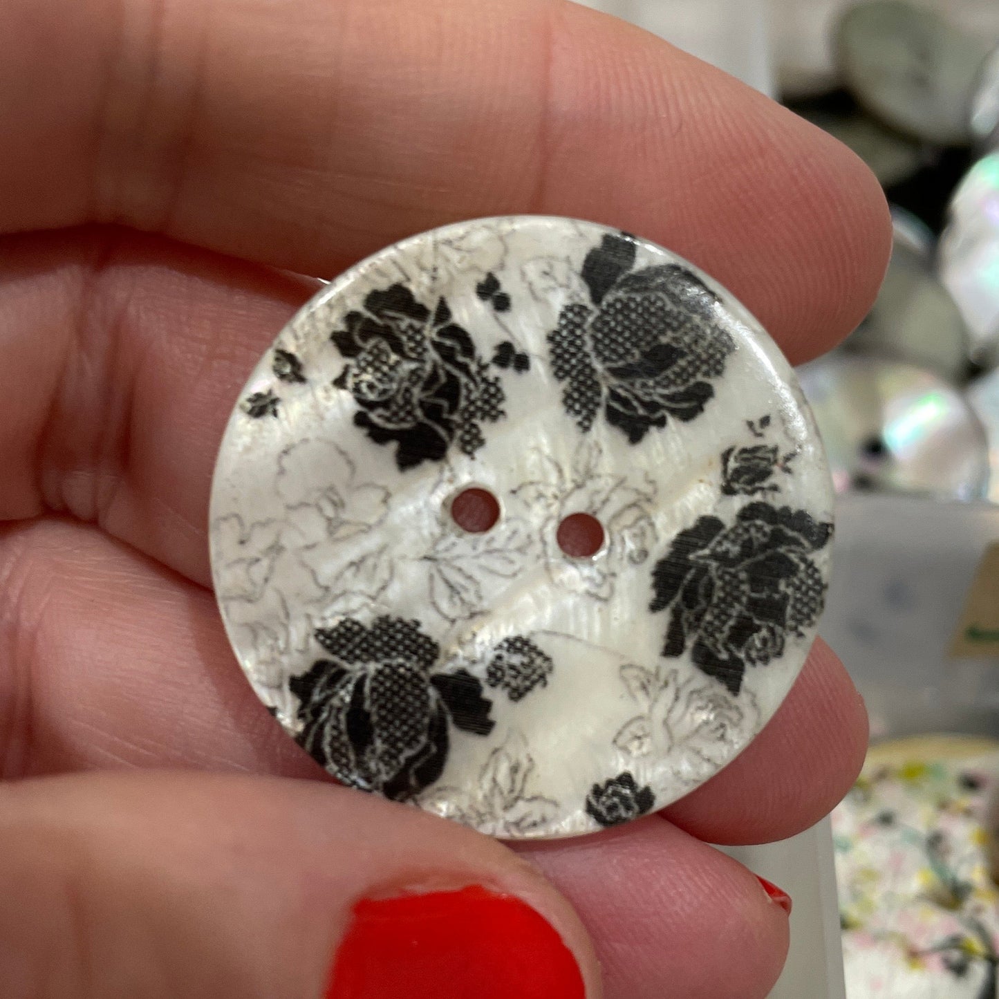 Hand holding a decorative button with black floral pattern against a blurred background of similar buttons.