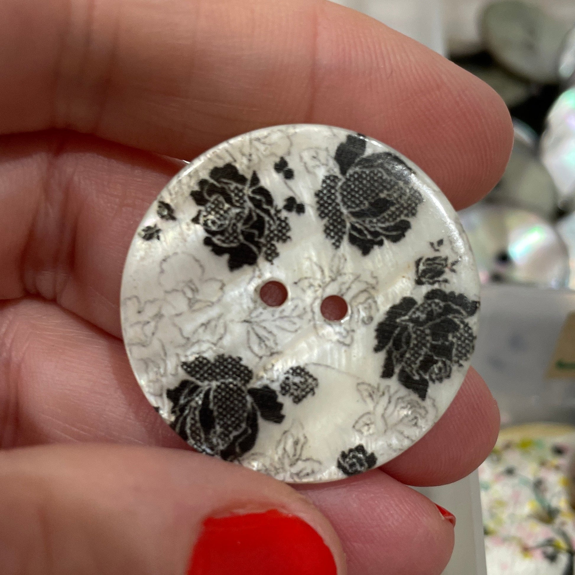 Hand holding a decorative button with black floral pattern against a blurred background of similar buttons.