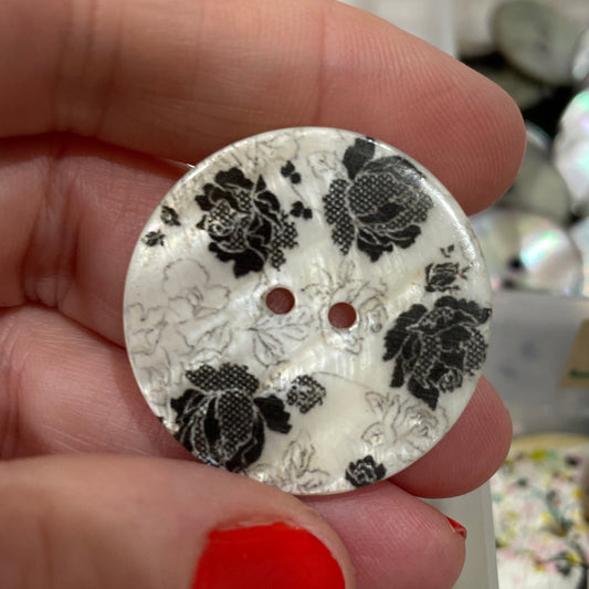Hand holding a decorative button with black floral pattern against a blurred background of similar buttons.
