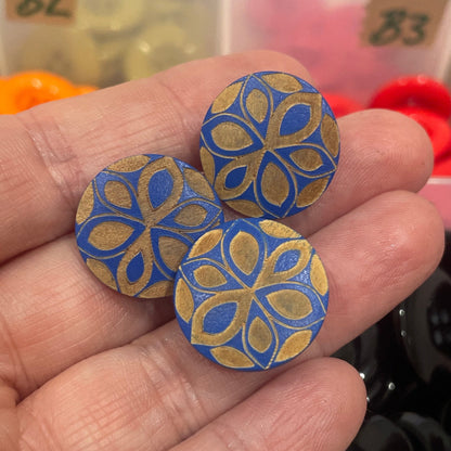 Hand holding two decorative buttons with a colorful pattern against a background of button storage.