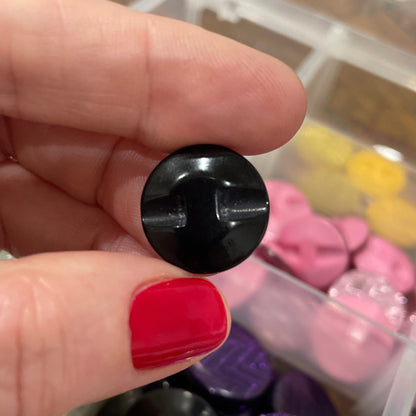 Black button held by a hand with red nail polish, against a background of colorful buttons in a container.