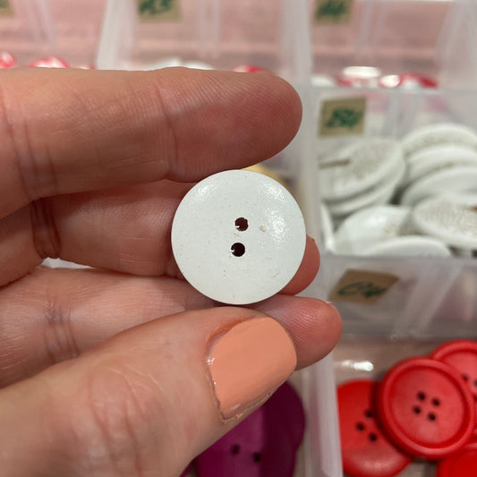 White button held between fingers with a background of various buttons in a box.