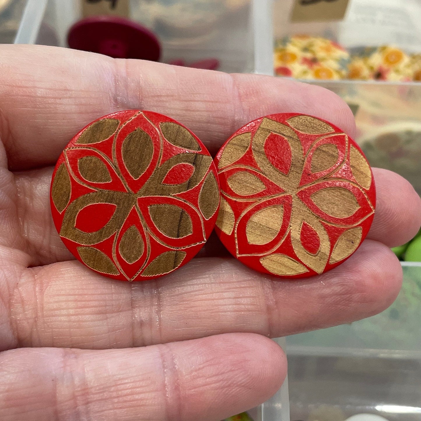 Hand holding two red floral-patterned buttons with a background of a button organizer.