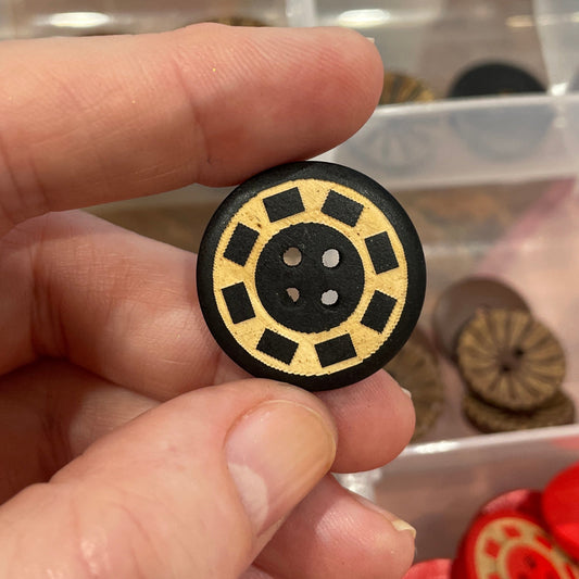 Hand holding a decorative button with a detailed design, surrounded by a collection of buttons in a clear organizer.