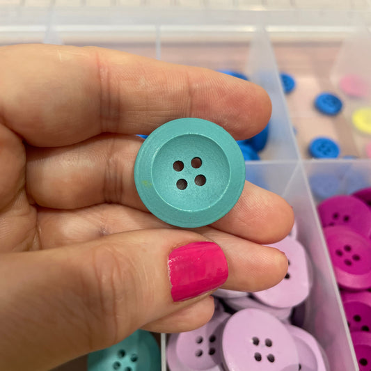 Hand holding a teal button with a container of colorful buttons in the background