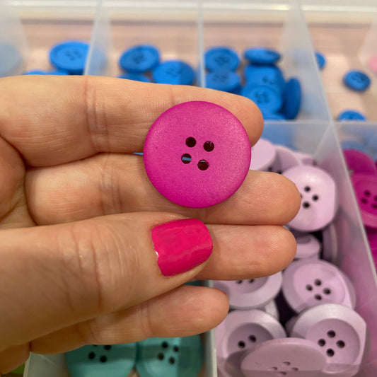 Hand holding a pink button with a container of buttons in the background