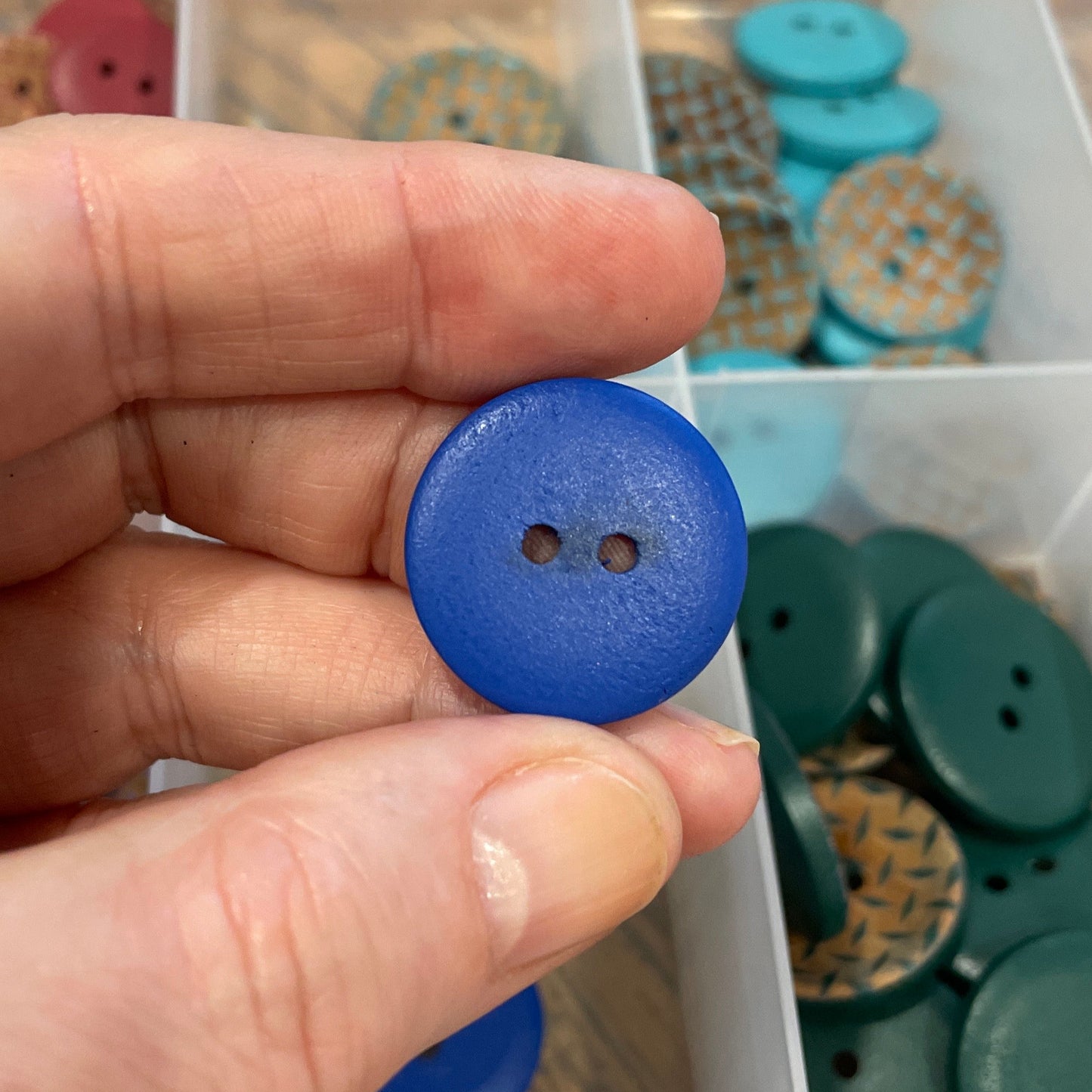 Hand holding a blue button with a container of assorted buttons in the background