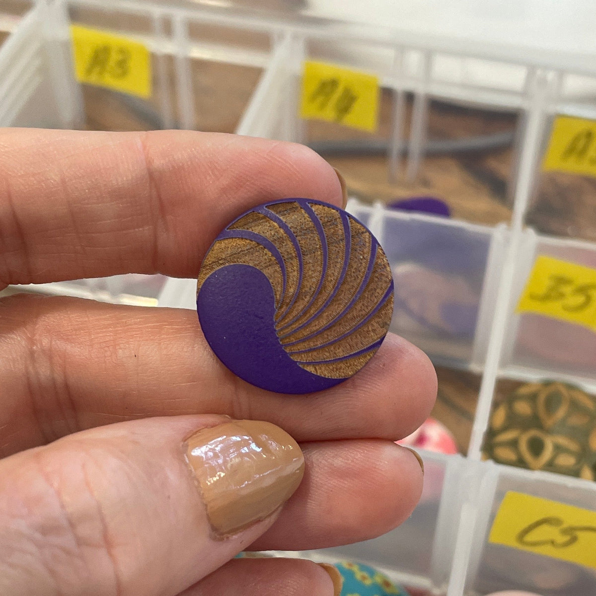 Hand holding a decorative button with a colorful design, set against a background of various buttons in labeled compartments.