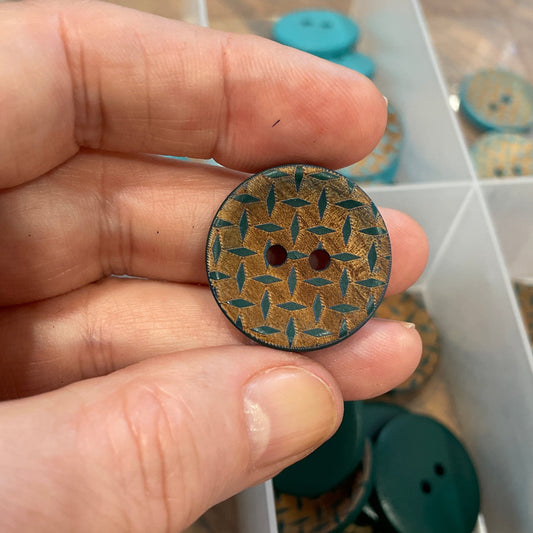 Hand holding a decorative button with a pattern, against a background of a clear container with more buttons.