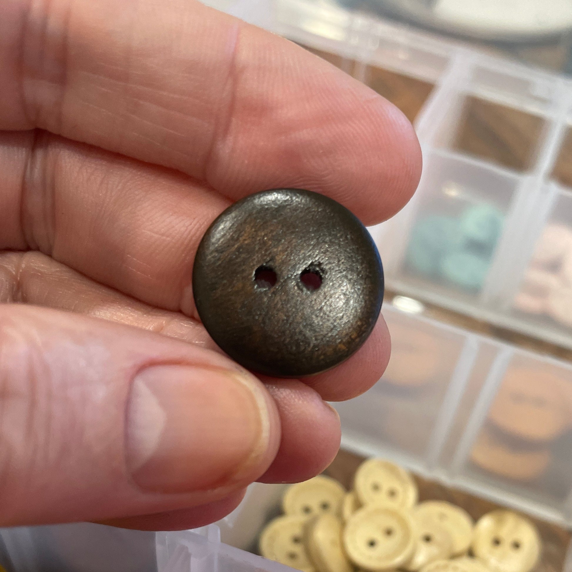 Hand holding a dark brown button with a background of colorful buttons in containers.