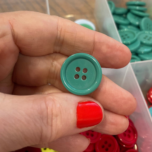 Hand holding a teal button with a container of colorful buttons in the background
