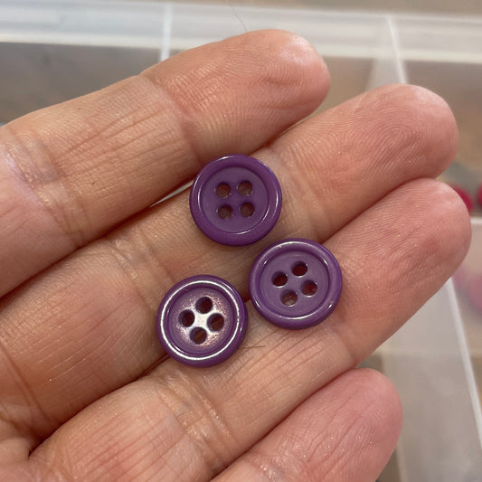 Hand holding three purple buttons with a background of button storage container.