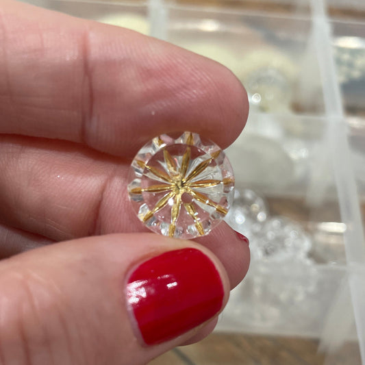 Gold and diamond earring held by a hand with red nail polish, against a clear jewelry box background.