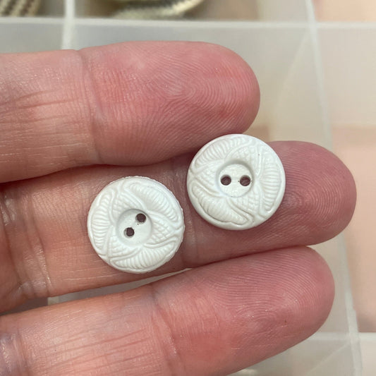 Two white buttons held between fingers with a background of various buttons in a box.