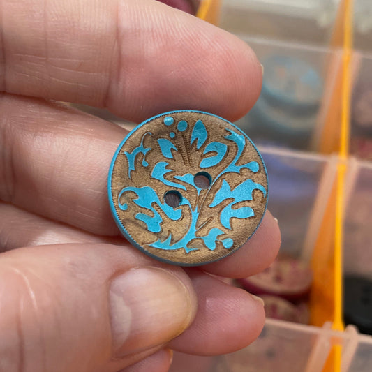 Hand holding a decorative button with intricate design, blurred button collection in the background