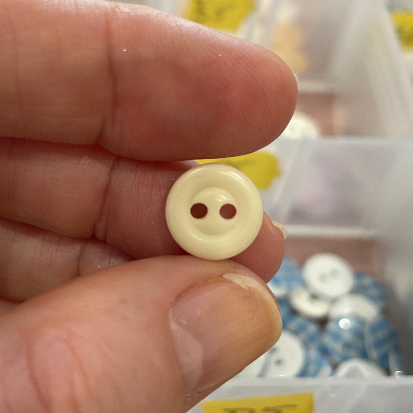 Hand holding a beige button with two holes in front of a container of buttons.