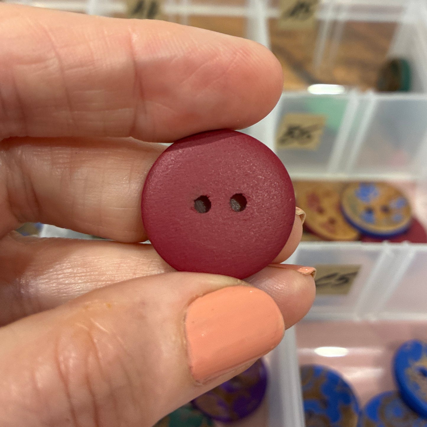 Hand holding a red button with two holes above a container of colorful buttons.