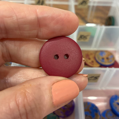 Hand holding a red button with two holes above a container of colorful buttons.
