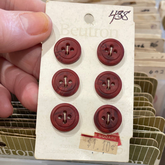 Set of six brown buttons on a card held by a hand, with a clear plastic container in the background.