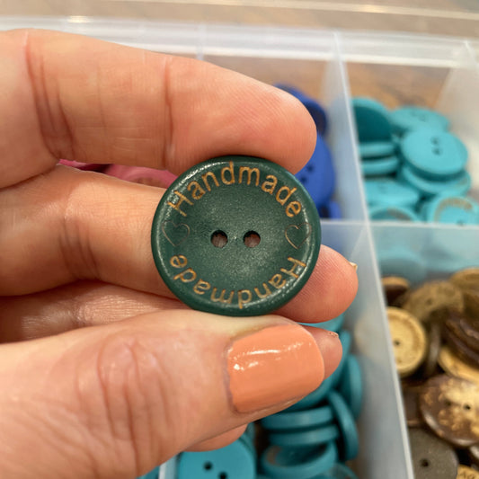 Hand holding a green button with 'Handmade' text, surrounded by blue buttons in a container.