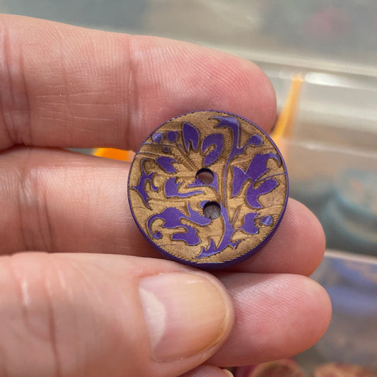 Hand holding a decorative button with a colorful design, set against a background of more buttons in a container.