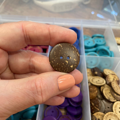 Hand holding a brown button with a container of assorted buttons in the background