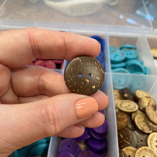 Hand holding a brown button with a container of assorted buttons in the background