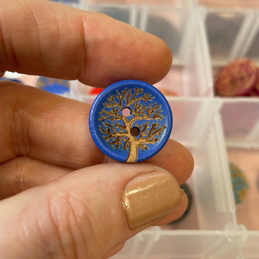 Hand holding a blue button with a tree design, with a background of more buttons in a container.