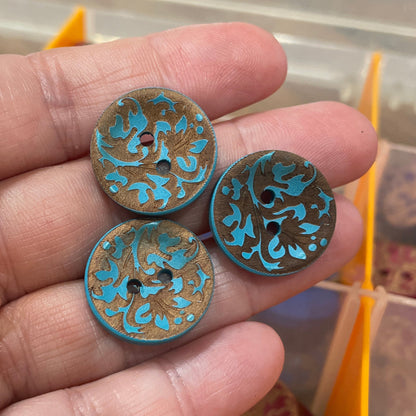 Three decorative buttons with blue and gold patterns held in a hand, with a container of more buttons in the background.