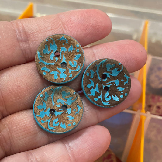 Three decorative buttons with blue and gold patterns held in a hand, with a container of more buttons in the background.