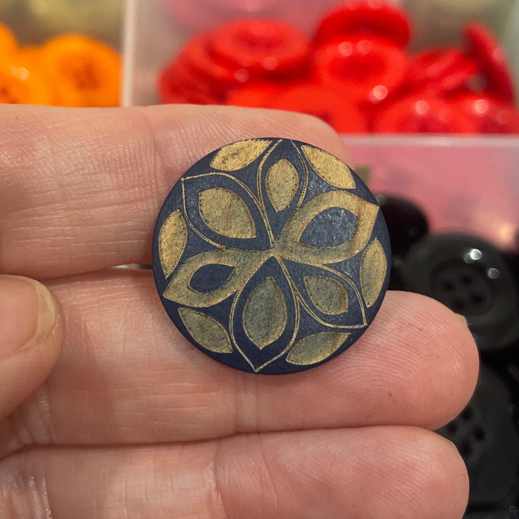 Hand holding a decorative button with a floral pattern against a background of colorful buttons in a box.