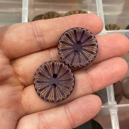 Hand holding two decorative buttons with a container of various buttons in the background
