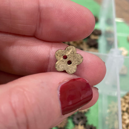Gold flower-shaped button  held between fingers with a container of similar buttons in the background.