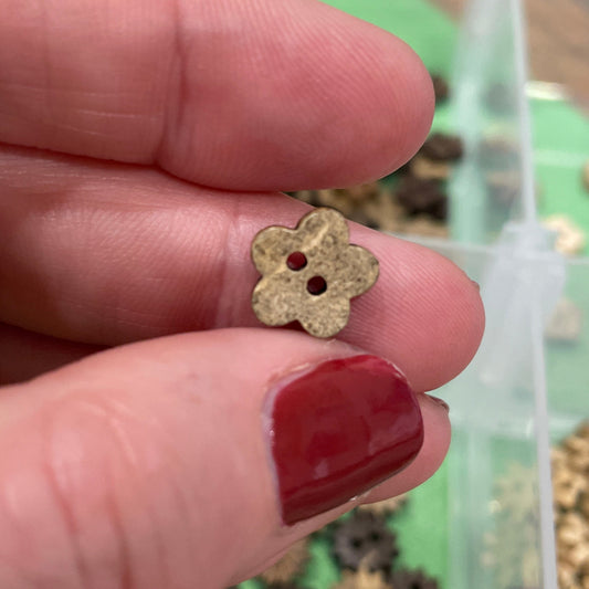 Gold flower-shaped button  held between fingers with a container of similar buttons in the background.