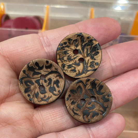 Hand holding three decorative buttons with a pattern, with a button collection container in the background.