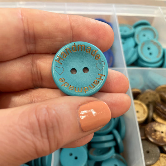 'Handmade' turquoise button held by a hand with a container of buttons in the background
