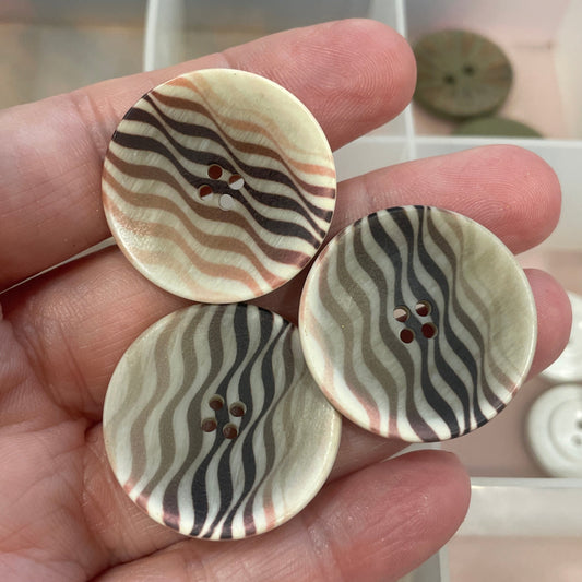 Three patterned buttons held in a hand with a background of more buttons in a container.