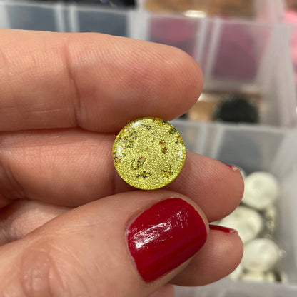 Gold glittery earring held between fingers with a background of assorted earrings in a box.