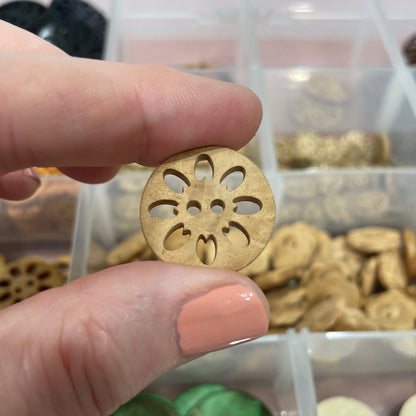 Hand holding a gold button with intricate design, surrounded by various buttons in a clear container.