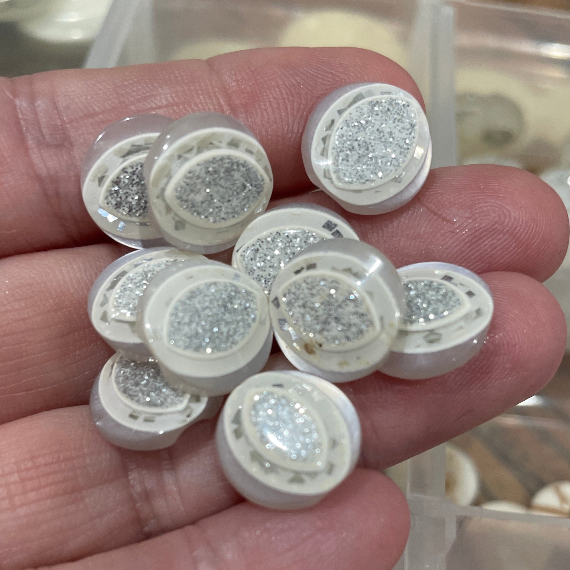 Hand holding small round decorative items with glittery surface, blurred container of similar items in the background.